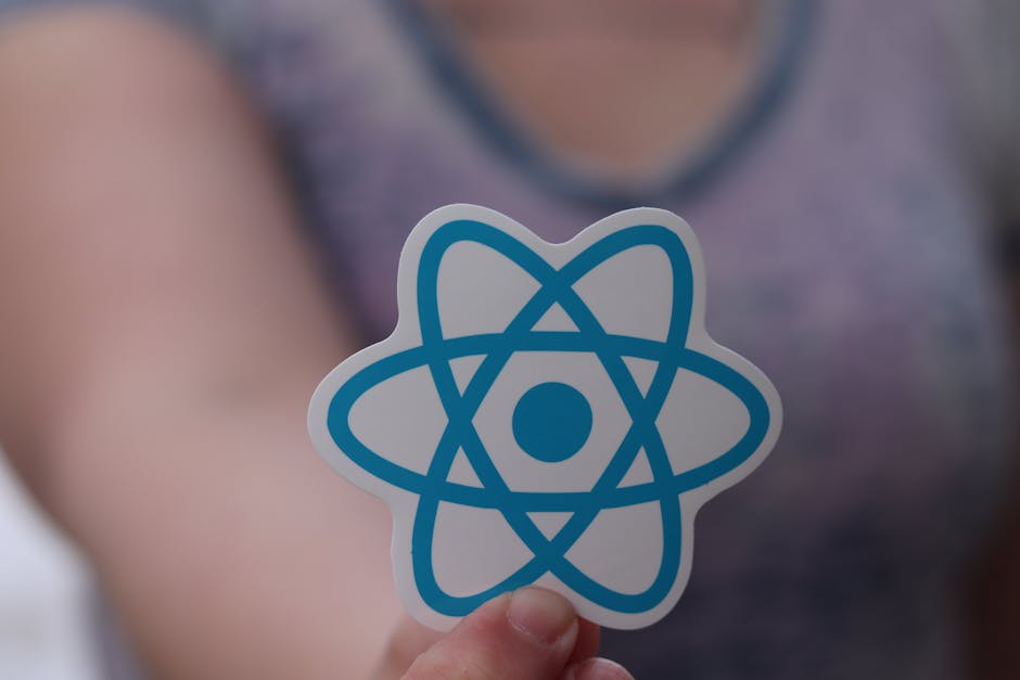 A person holding a paper cutout of an atom symbol, featuring blue orbitals and a central dot, against a blurred background that appears to be part of a casual indoor setting with a person wearing a gr