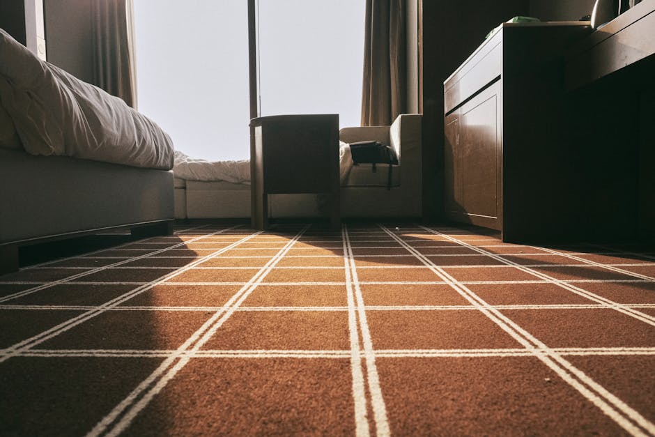 A person using a yellow vacuum cleaner with a black hose attachment to clean a traditional patterned carpet in a room. The individual is wearing beige shoes, grey pants, and a beige jacket, and is sta
