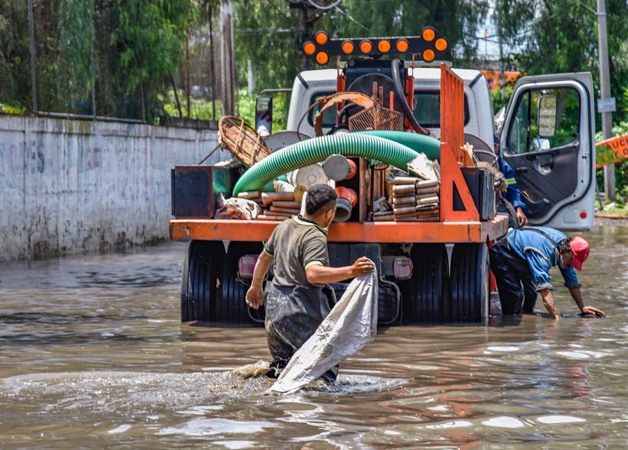 Two men are working to clear floodwaters near a large orange and white utility truck loaded with various tools and equipment, parked on a flooded street with water reaching mid-calf. One man, wearing 