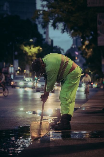 Two men are working to clear floodwaters near a large orange and white utility truck loaded with various tools and equipment, parked on a flooded street with water reaching mid-calf. One man, wearing 