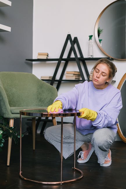 A young woman wearing a lavender sweatshirt and yellow cleaning gloves is kneeling on a dark wooden floor, polishing a round glass-topped side table with a pink cloth. The room features a light grey w